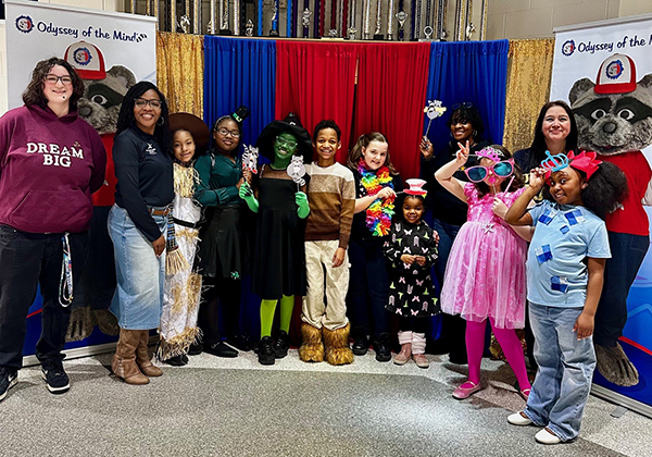 A group of students, and adults pose in front of a colorful red, blue, and gold curtain backdrop at an Odyssey of the Mind event. Several children are dressed in costumes including a Wicked Witch, Scarecrow, and other characters, while others hold fun photo booth props. Odyssey of the Mind banners with the raccoon mascot are visible on both sides.