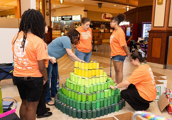 NNPS students in orange shirts stack canned goods into a pyramid in a mall.