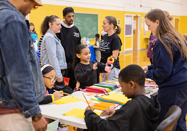 Students sitting at a table drawing with parents and teachers standing around them.