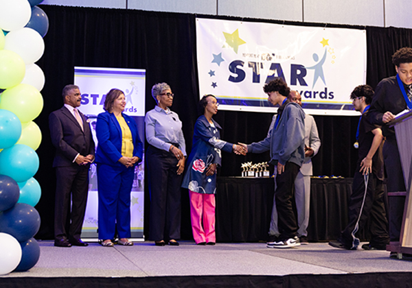 A student shakes hands with Superintendent Dr. Michele Mitchell on stage at the Newport News Public Schools STAR Awards ceremony, while other district leaders stand nearby. Gold star trophies are visible on a table behind them, and a colorful balloon arch and STAR Awards banner decorate the stage.

.