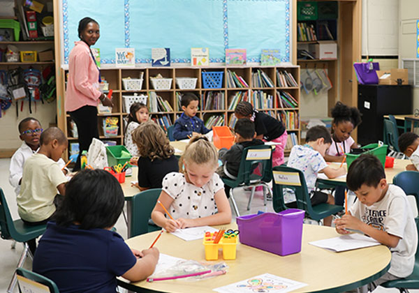 Elementary students working at round tables in a classroom while a teacher stands nearby
